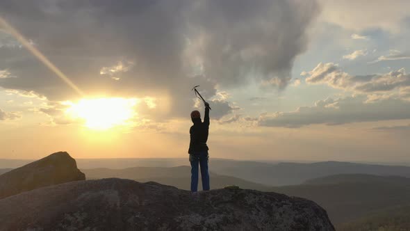 Drone Shot of a Man Standing on Top of a Rock Mountain and Joyfully Waving His Hands Holding an Ice