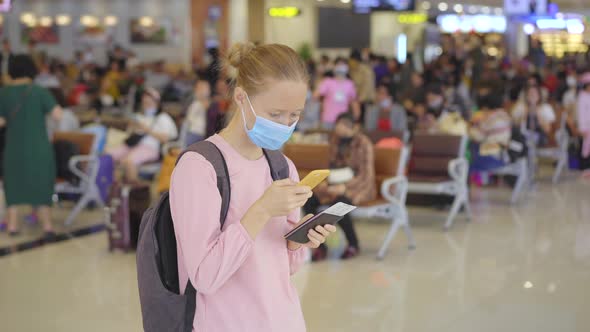 A Young Woman Wearing a Medical Face Mask in an Airport Stands in a Hall with a Cellphone in Her alt