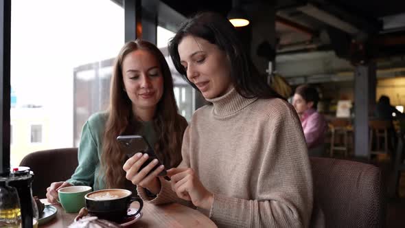 Two Young Attractive Girls in a Cafe are Looking at the Phone