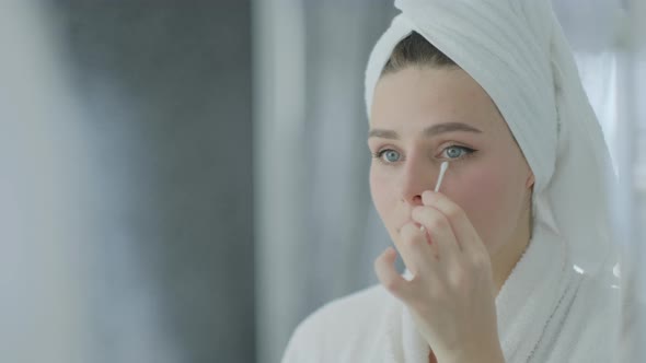 Attractive brunette woman removing make up with cotton stick and looking at reflection in mirror alt