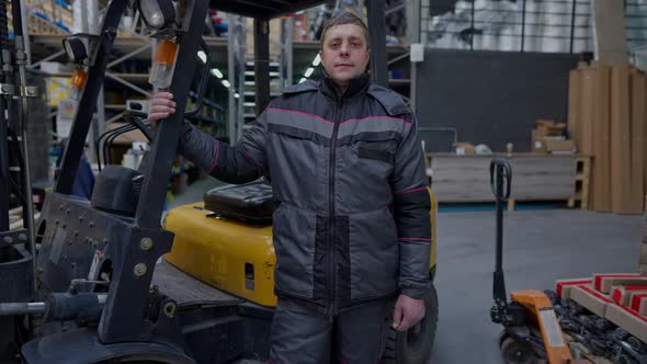 Zoom in to Confident Adult Man in Uniform Standing at Forklift Autoloader Looking at Camera alt