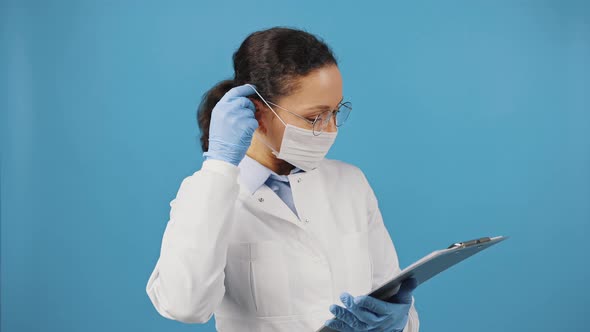 Woman Doctor with Clipboard Putting on Protective Medical Mask Before Appointment with Patient alt