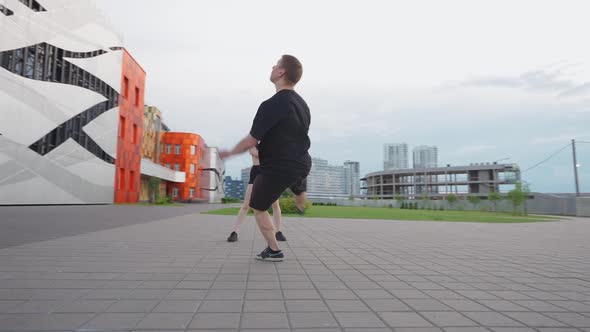 Young Man Doing a Somersaults on Training Area Near Modern Buildings Guy and a Girl Perform alt
