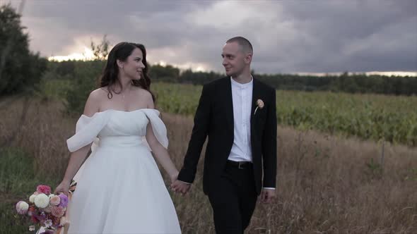 The Bride with a Bouquet of Dahlias Holds the Groom's Hand and They Walk Across the Field Looking alt