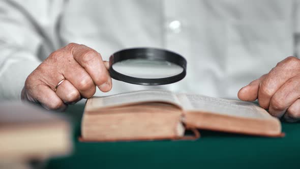 Closeup Elderly Wrinkled Male Hands Reading Antique Paper Book Use Magnifying Glass at Table Home alt