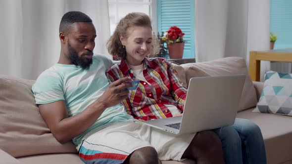 Happy Multiethnic Couple Shopping Online with Credit Card and Laptop at Home alt