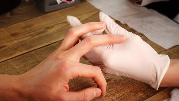 Nail Master in White Gloves Makes Manicure to Male Client Using a File and Brush in Closeup alt