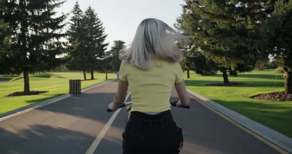 Beautiful Blonde Girl Teenager Rides Bicycle in Park, View From the Back