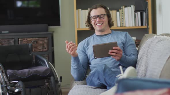 Happy caucasian disabled man sitting on sofa in living room using tablet alt