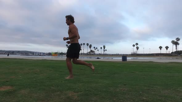 Tracking shot of a man jogging on on grass near a bay alt