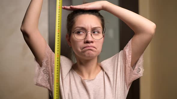 A young woman shows sadness at her height by holding a measuring tape alt