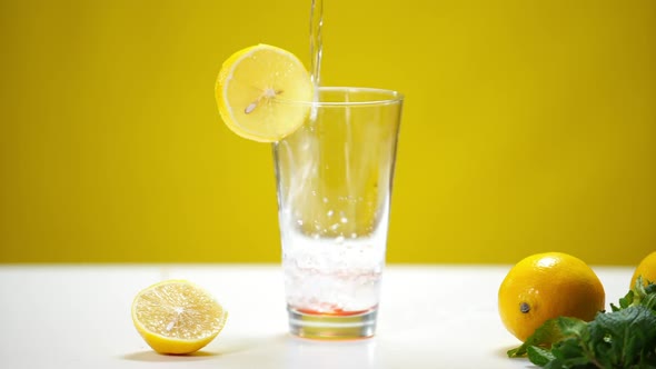 Glass with Lemon Slice at Yellow Background and Water Flowing Inside Spilling alt