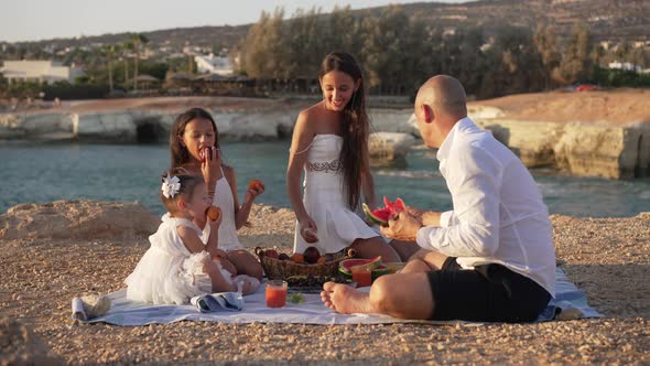 Wide Shot of Happy Family Eating Delicious Vitamin Fruits in Sunshine of Golden Sunset on Sandy Sea alt