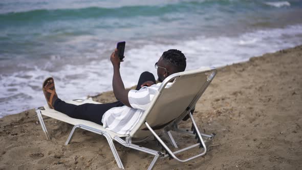 Carefree African American Man Taking Selfie on Smartphone Lying on Sunbed with Azure Blue Waves alt