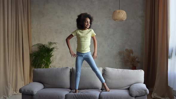 Portrait Little Cheerful African American Girl Dancing and Jumping on the Couch alt