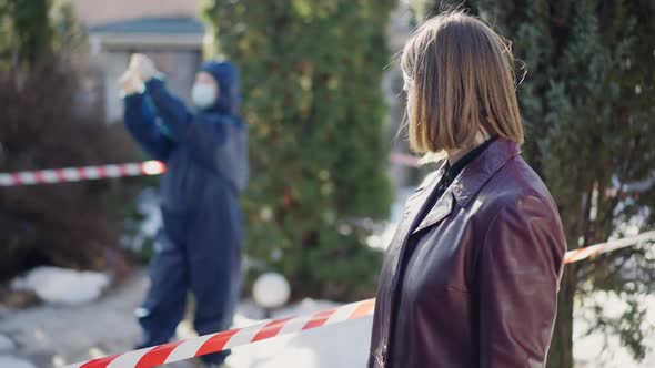 Young Female Detective Looking Back at Blurred Criminalist Gathering Clues and Looking at Camera alt