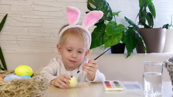 A little boy in rabbit ears paints Easter eggs with paint and smiles