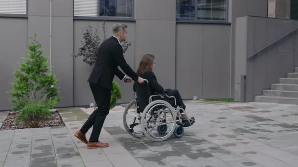 Man in Suit Pushing Wheelchair with Woman Coworker To Office Building Stairs alt