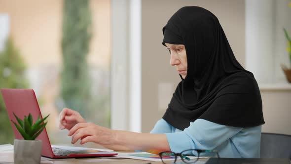 Side View Portrait of Intelligent Middle Eastern Mature Woman in Hijab Messaging Online on Laptop alt