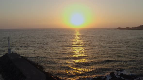 A breakwater and a small lighthouse at sunset. A drone flying towards the sun. alt