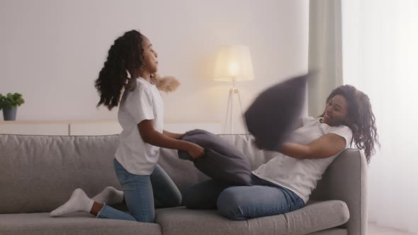 Happy African American Mother and Daughter Playing Pillow Fight Falling Down on Sofa Hugging and alt