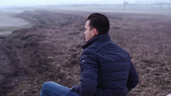 Pensive Man Sits on Bare Beach Stones at Vast Grey Sea alt