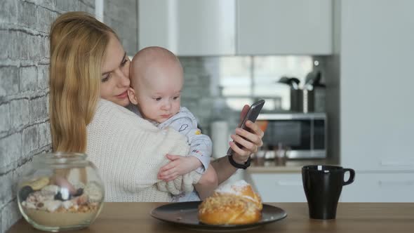 mom with baby in her arms uses a smartphone while sitting in the kitchen