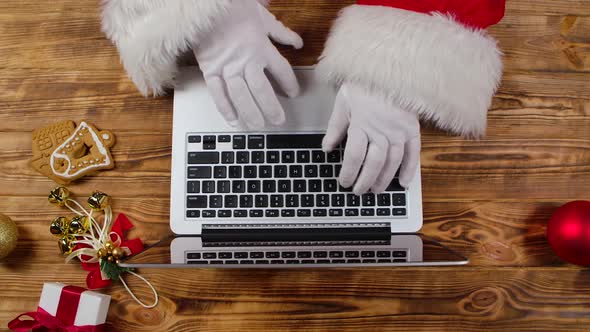 Top View Santa Hands in White Gloves Typing on the Keyboard By Wooden New Year Decorated Table alt