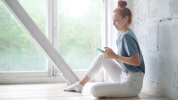 Happy Redhead Young Woman Wearing Wireless Earphones Is Using Cell Phone Sitting By the Big Window alt