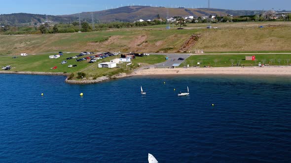 people practicing in the sailing school of the lake with beach, car parking and wind turbines in the alt