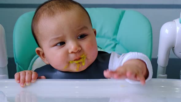 A dirty, joyful baby at the table on a feeding chair got his mouth dirty in vegetable puree and knoc alt