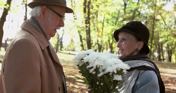 Happy Elder Woman Sniffs a Bouquet of Flowers and Rejoices with Her Man in Park alt