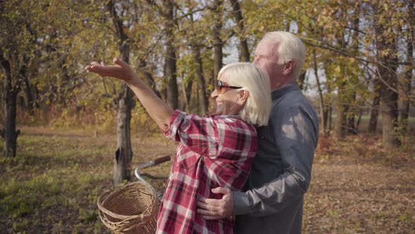 Happy Caucasian Retired Couple Standing in Sunlight in the Autumn Forest or Park and Smiling alt