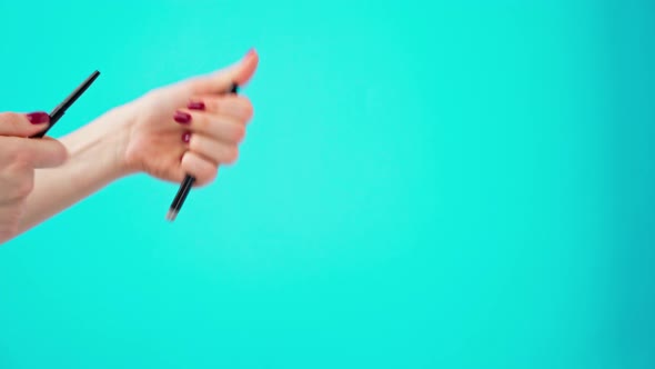 Female Hands Holding Cosmetic Eye Liner Pencils Against Blue Background alt