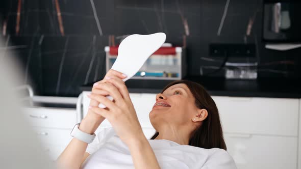 A Patient with Braces Looks Intently in the Mirror After Installing Braces Sitting in a Dental Chair alt