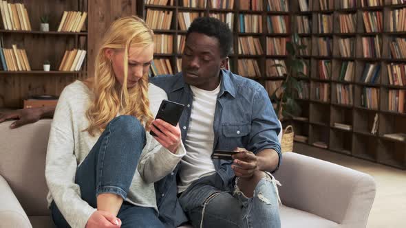 Mixed Race Couple Sit on Sofa Use Smartphone and Credit Card Make Order Buying on Internet Laughing alt