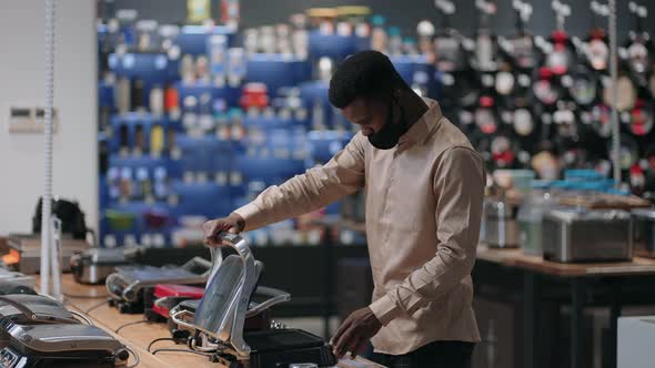 Young Black Man with Face Mask is Shopping in Home Appliances Store Viewing Samples of Electric alt