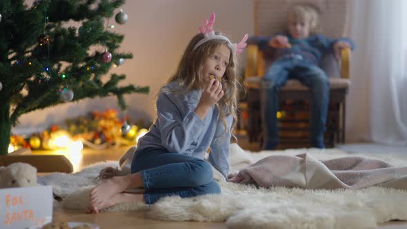 Wide Shot Portrait Charming Little Thoughtful Girl Eating Christmas Cookie with Boy in Rocking Chair alt