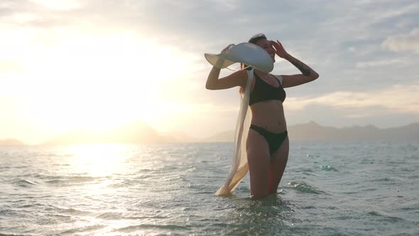 Woman In Bikini Holding Sun Hat Standing In Sea At Sunset alt