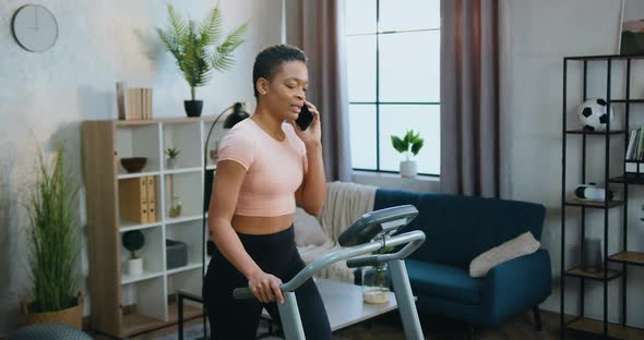 Woman in Training Clothes Walking on Treadmill During Home Morning Workout and Talking on Phone alt