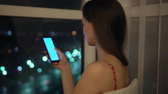 girl holds phone and looks at it at window against background of night city. alt