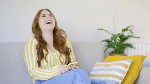 Young woman sitting on sofa laughing about text message on smartphone