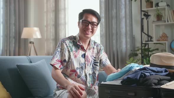 Asian Man Packing Clothes In Suitcase And Warmly Smile To Camera At Home, Preparing For Vacation alt