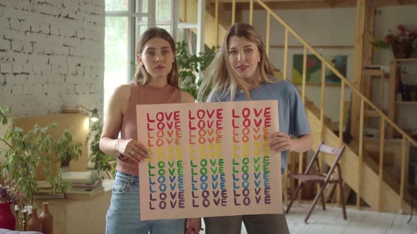 Two Attractive Young Girls are Standing in the Studio and Showing a Lgbt Poster at the Camera alt