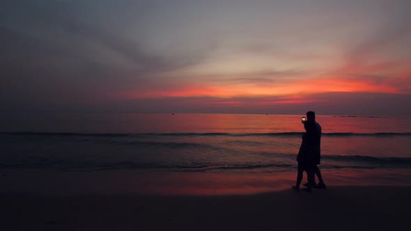 Cinematic View of Young Couple Walking at the Beach and Taking Photos at Dusk alt