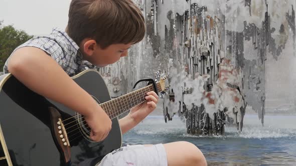 Portrait A Cute Caucasian Boy Sings a Song While Playing an Acoustic Guitar Near a Water Fountain in