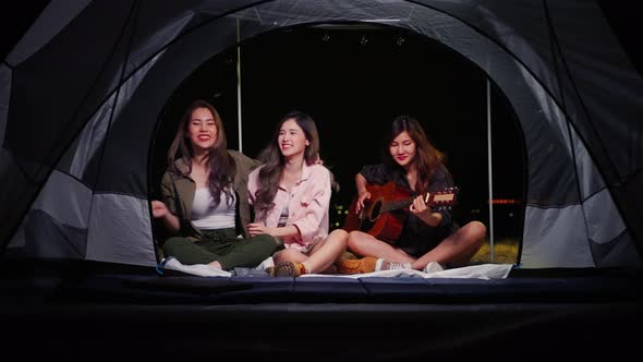 group of young Asian friends playing guitar and singing in a tent during a summer camping