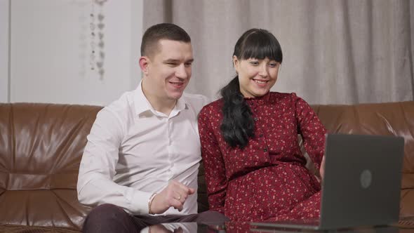 Happy Pregnant Caucasian Couple Waving at Laptop Talking and Caressing Belly alt