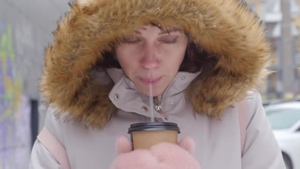 A Young Girl Drinks Tea Through a Straw on a City Street in Winter