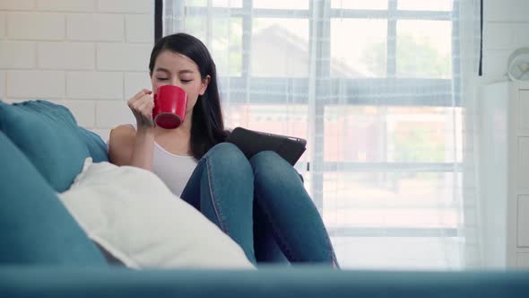 Asian woman using tablet holding a warm cup of coffee or tea while lying on the sofa. alt